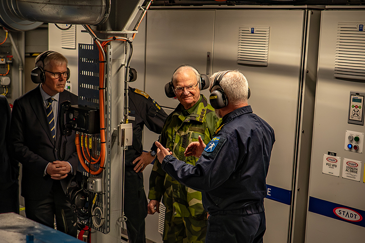 Kungen i samspråk med andra i maskinrum. Hörlurar på.