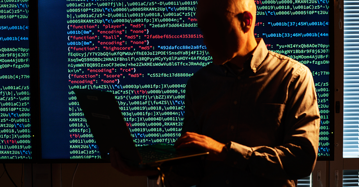 Cyberförsvarare med en bärbar dator i ett mörkt rum framför en bildskärm med datakod. Klicka på bilden för att få den förstorad.